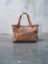 Încărcați imaginea în vizualizatorul Galeriei, Geantă shopper maro cu bretele din piele pe podea de beton, cu perete gri pe fundal.