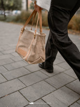 Încărcați imaginea în vizualizatorul Galeriei, Persoană care ține o geantă shopper bej cu mânere maro pe o alee pavată.