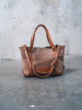 Load image into Gallery viewer, Geantă maro din piele pe o suprafață gri, cu perete de beton pe fundal.

