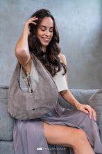 Load image into Gallery viewer, Femeie care ține o geantă gri, așezată pe o canapea, cu fundal neutru.

