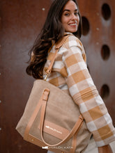 Load image into Gallery viewer, Femeie care ține o geantă bej, purtând o jachetă în carouri, pe un fundal maro texturat.

