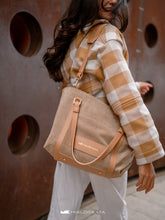 Load image into Gallery viewer, Persoană care poartă o geanta bej cu bretele maro, pe fundal cu perete texturat.

