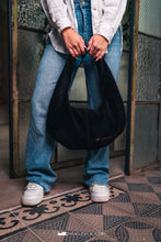 Load image into Gallery viewer, Persoană care ține o geantă neagră, cu podea cu model și ușă metalică în fundal.

