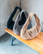 Load image into Gallery viewer, Trei genți din piele întoarsă în negru, gri și bej, așezate pe o bancă de lemn, pe fundal cu perete de beton.

