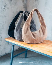 Load image into Gallery viewer, Trei genți din piele întoarsă în negru, gri și bej, așezate pe o bancă de lemn, pe fundal cu perete texturat.

