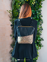 Kép betöltése a galériamegjelenítőbe: Persoană care poartă un rucsac negru cu detaliu auriu decorativ, pe fundal verde cu frunziș.

